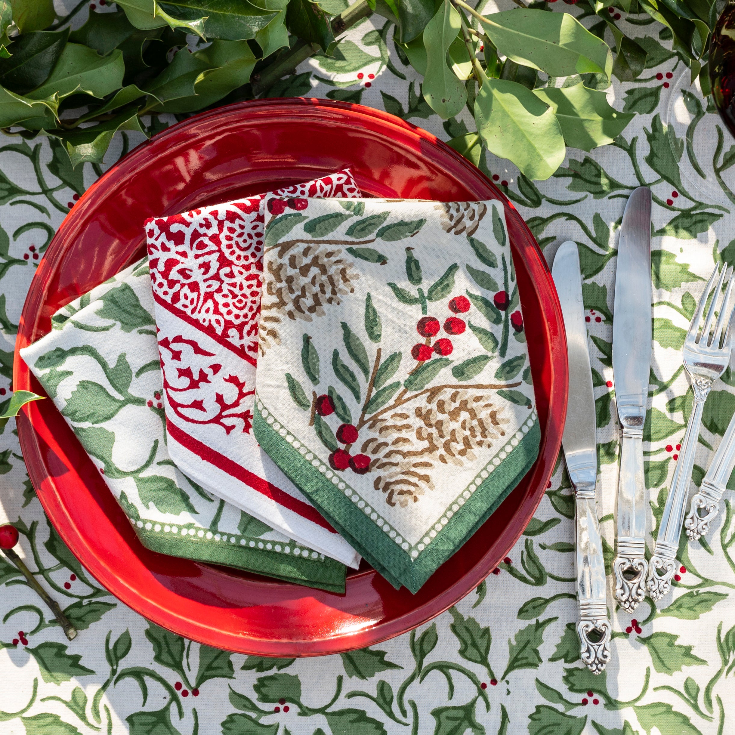 Green and white printed tablecloth layered with a red dinner plate and holly berry printed napkins and cutlery