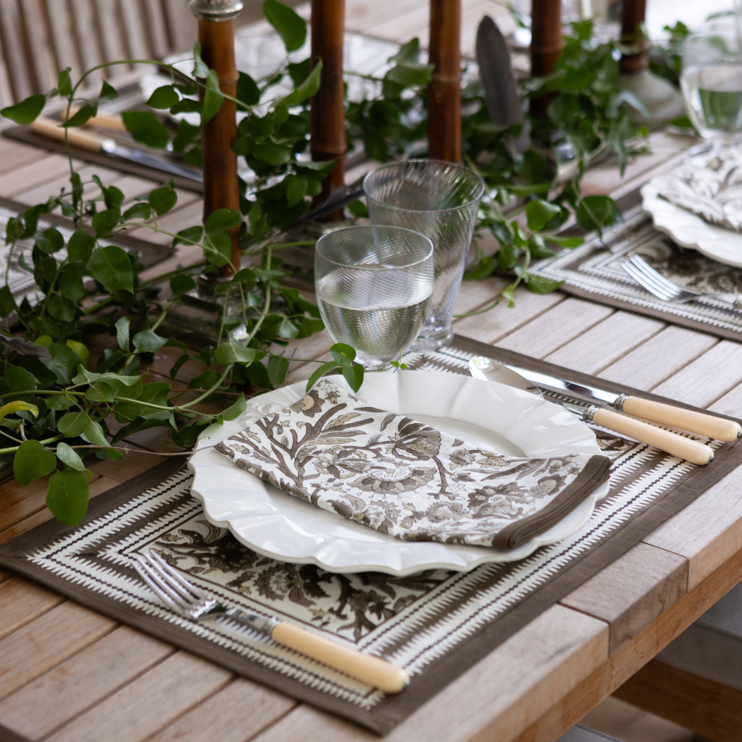 Dining table setting with greenery, plates, and cutlery on a wooden table.