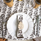 Dining table setting with white plates, silverware, and a leaf-themed napkin on a patterned tablecloth.