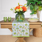 A cotton table runner with a tropical palm and pineapple pattern in light blue and green, displayed on a table with a vase of flowers, plates, and cutlery.