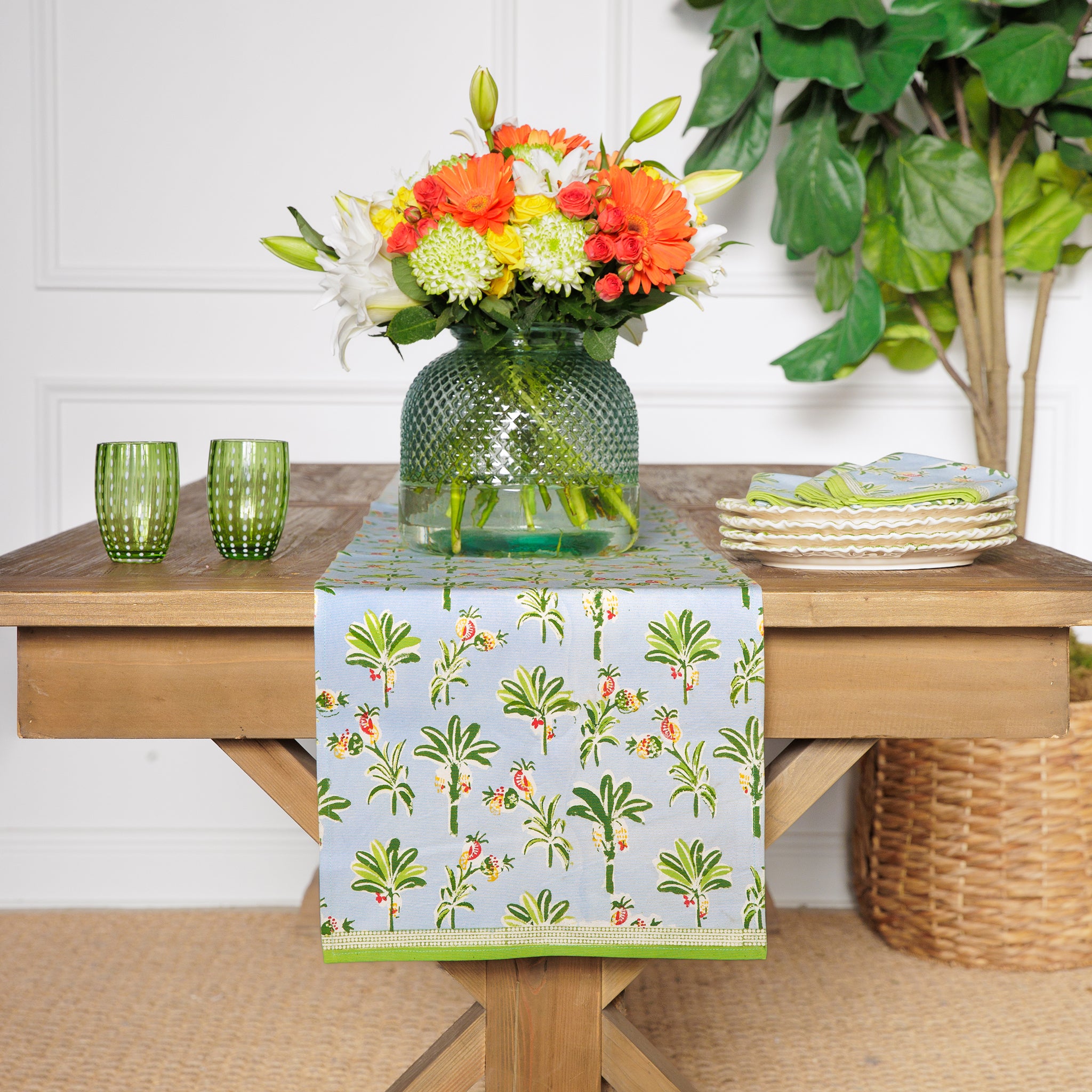 A cotton table runner with a tropical palm and pineapple pattern in light blue and green, displayed on a table with a vase of flowers, plates, and cutlery.