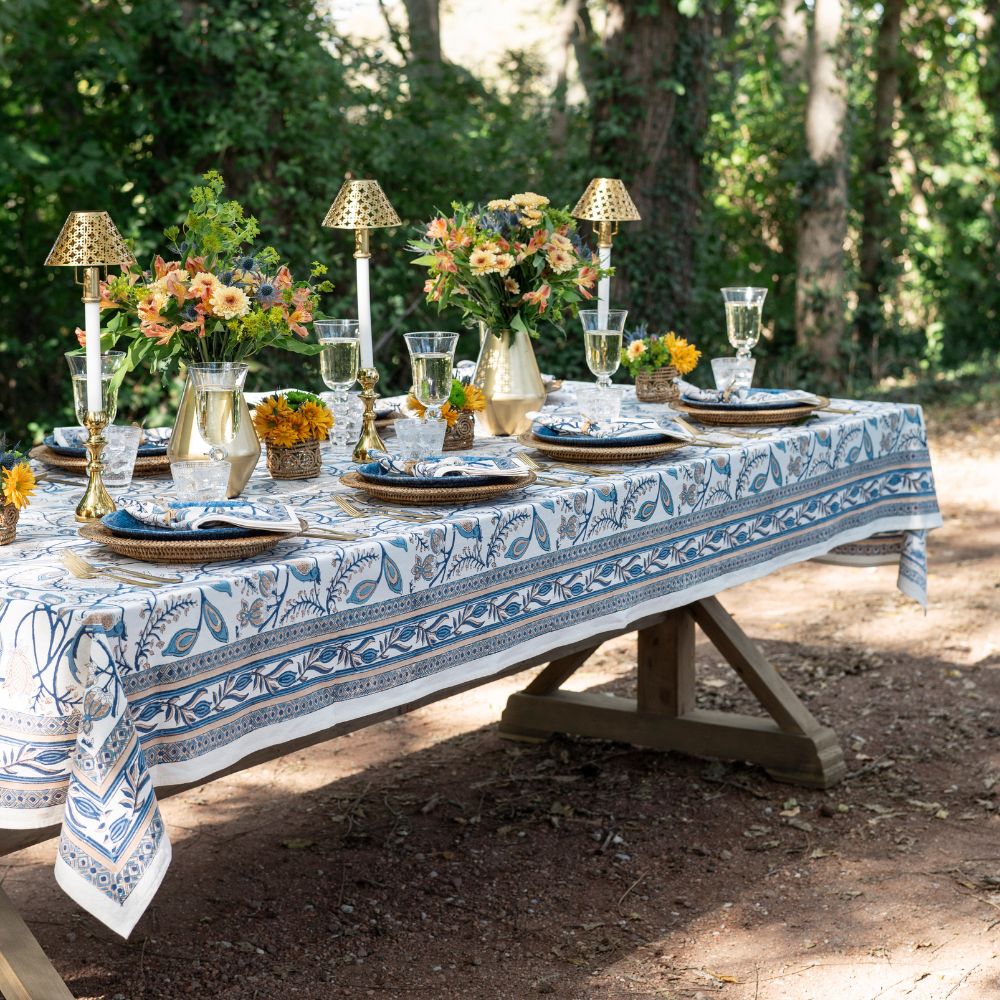 Outdoor table setting with a floral tablecloth, plates, glasses, and centerpieces in a natural setting.