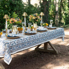 Outdoor table setting with a floral tablecloth, plates, glasses, and centerpieces in a natural setting.