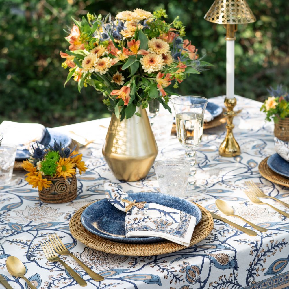 Elegant outdoor table setting with floral arrangements, gold vases, and blue plates.