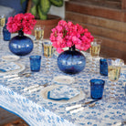A blue and white tablecloth with a floral pattern on a table, accompanied by blue and white dishes, blue glasses, and a blue vase with pink flowers.