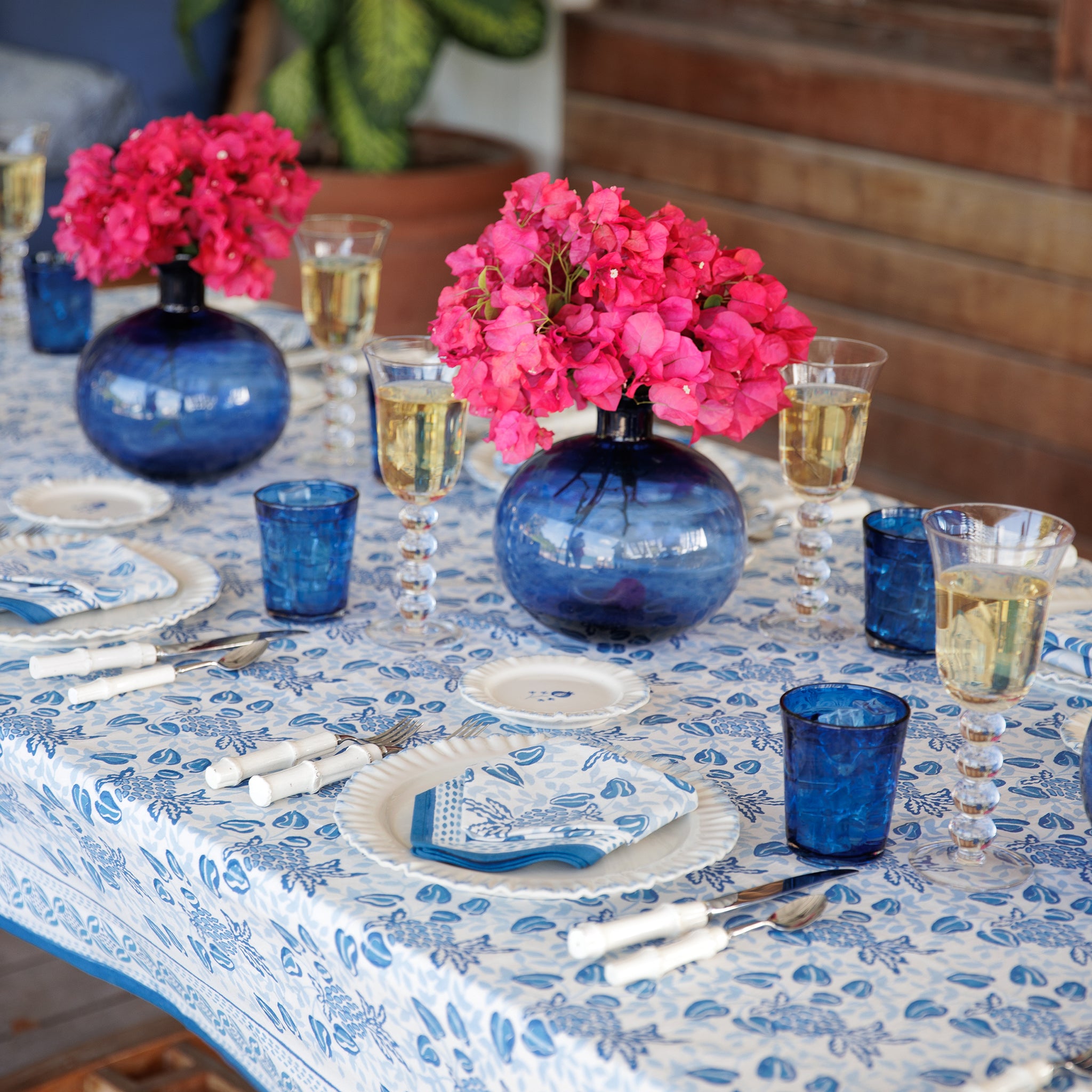 A blue and white tablecloth with a floral pattern on a table, accompanied by blue and white dishes, blue glasses, and a blue vase with pink flowers.