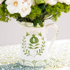 White ceramic pitcher with green floral patterns holding flowers on a decorative tablecloth.
