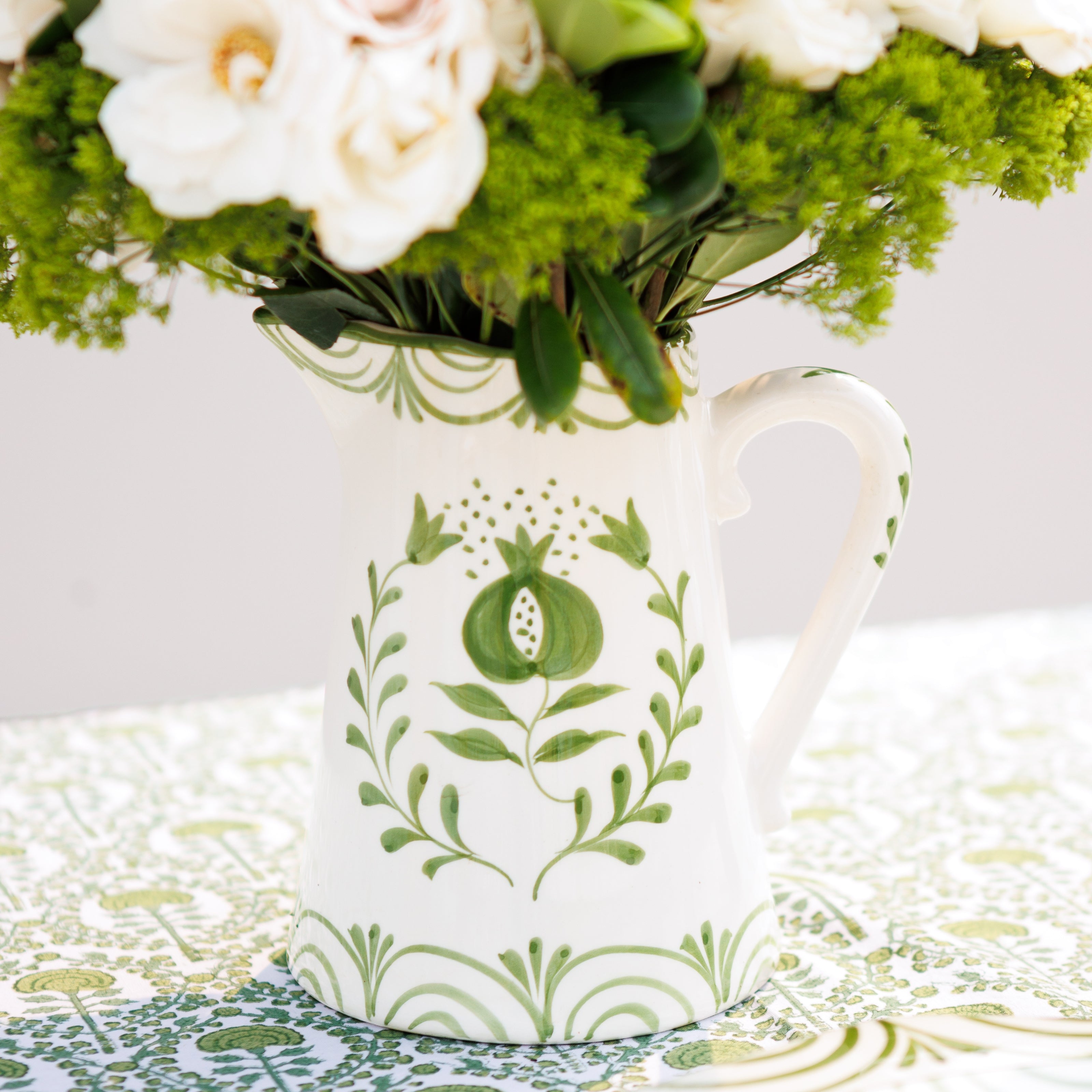 White ceramic pitcher with green floral patterns holding flowers on a decorative tablecloth.