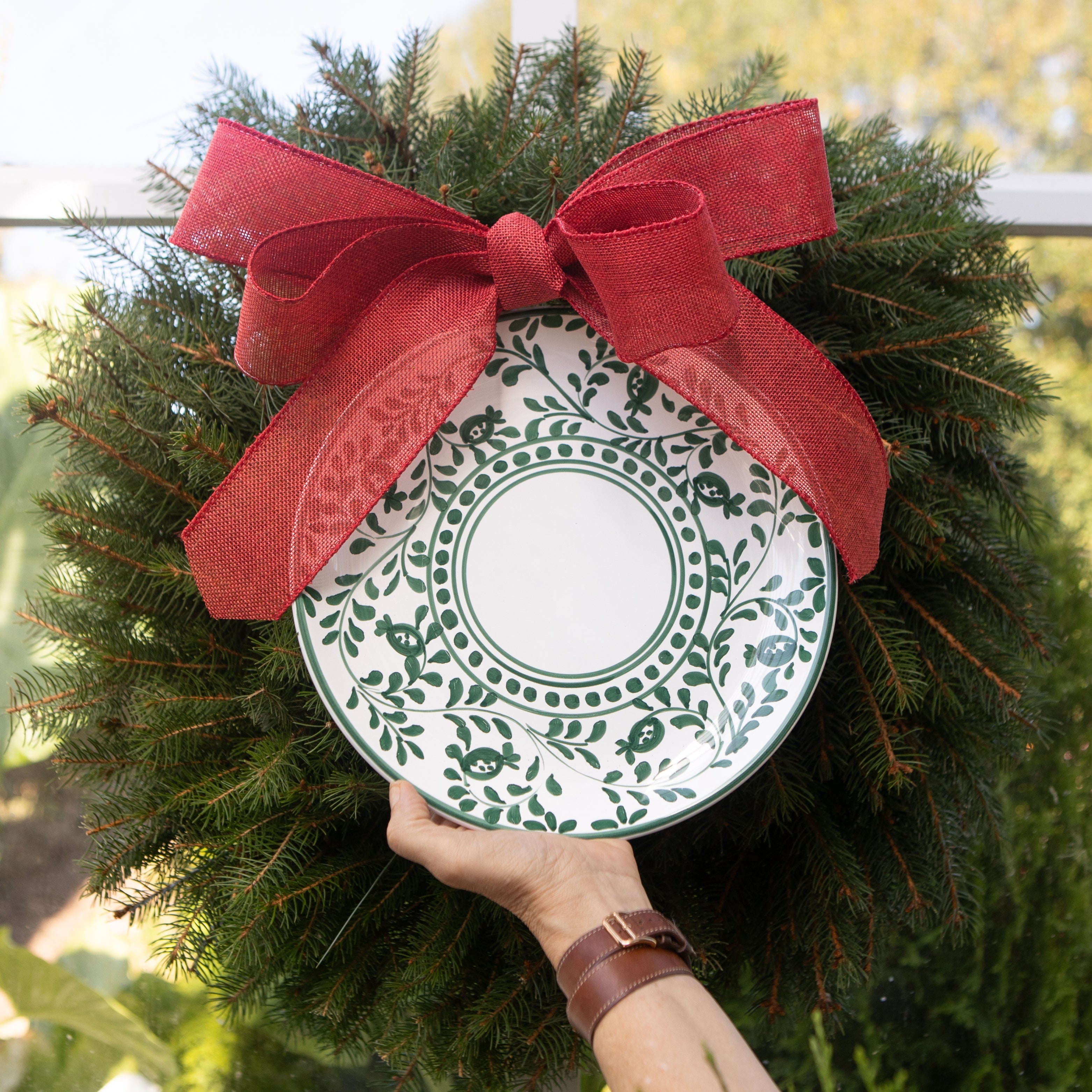 Decorative wreath with a green and white plate and red bow held by a hand.