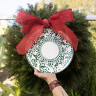 Decorative wreath with a green and white plate and red bow held by a hand.