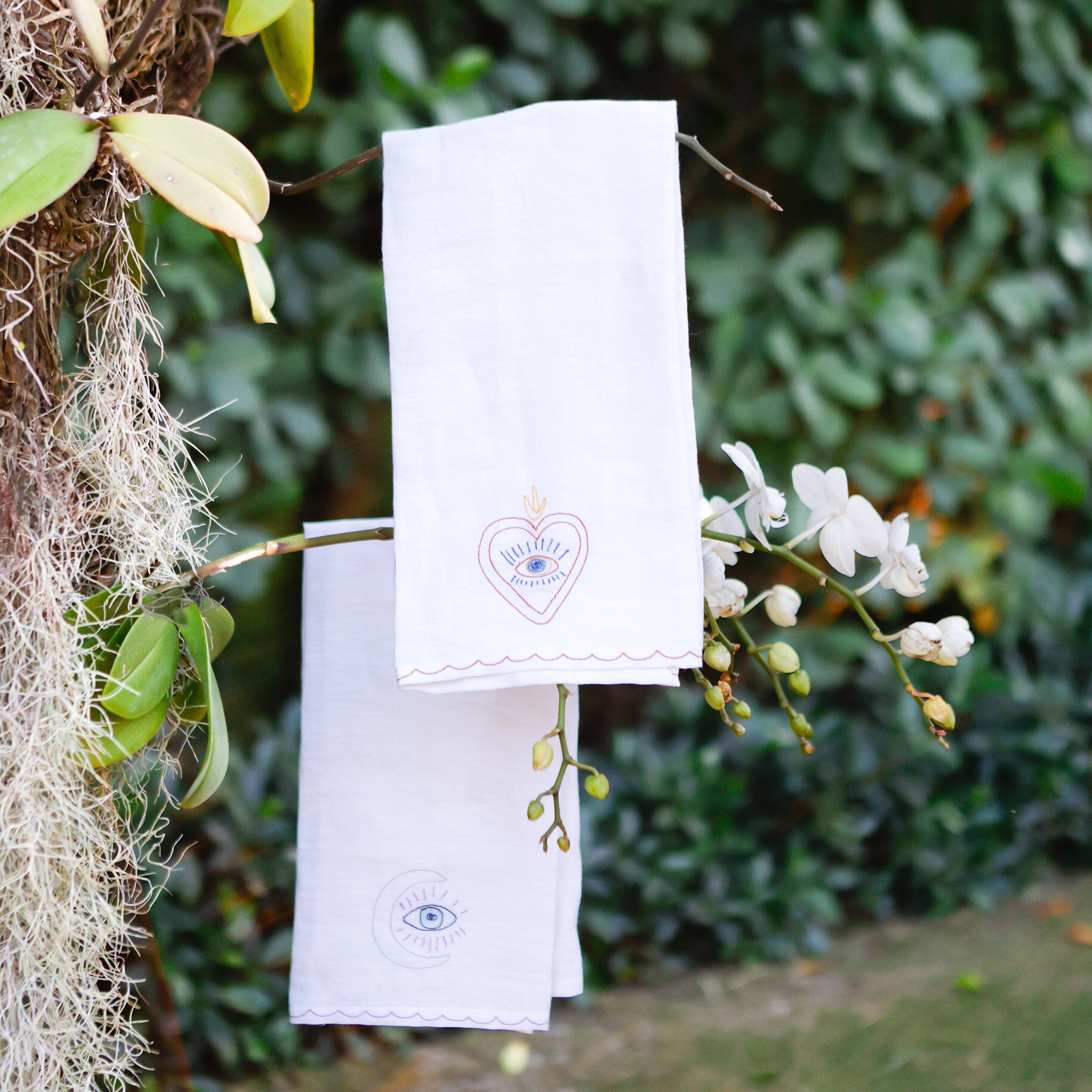 Two linen tea towels with embroidered designs, one featuring a heart with an eye symbol and the other featuring a crescent moon and star with an eye symbol, hanging on a rod with a greenery background.