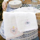 Two linen tea towels with embroidered designs, one featuring a heart with an eye symbol and the other featuring a crescent moon and star with an eye symbol, displayed on a woven tray