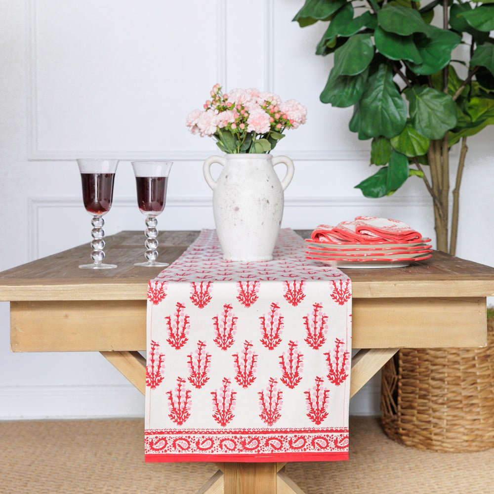 A red and white floral patterned table runner is displayed on a wooden table with a vase of flowers and a plant in the background.