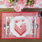 A red and white placemat with a floral pattern and paisley border, set on a wooden table with a white plate, red napkin, and silverware.