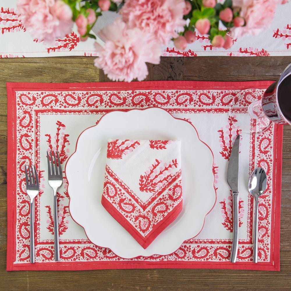 A red and white placemat with a floral pattern and paisley border, set on a wooden table with a white plate, red napkin, and silverware.
