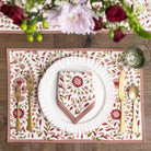 Table setting with floral placemat, white plate, gold cutlery, and pink napkin on a wooden table.