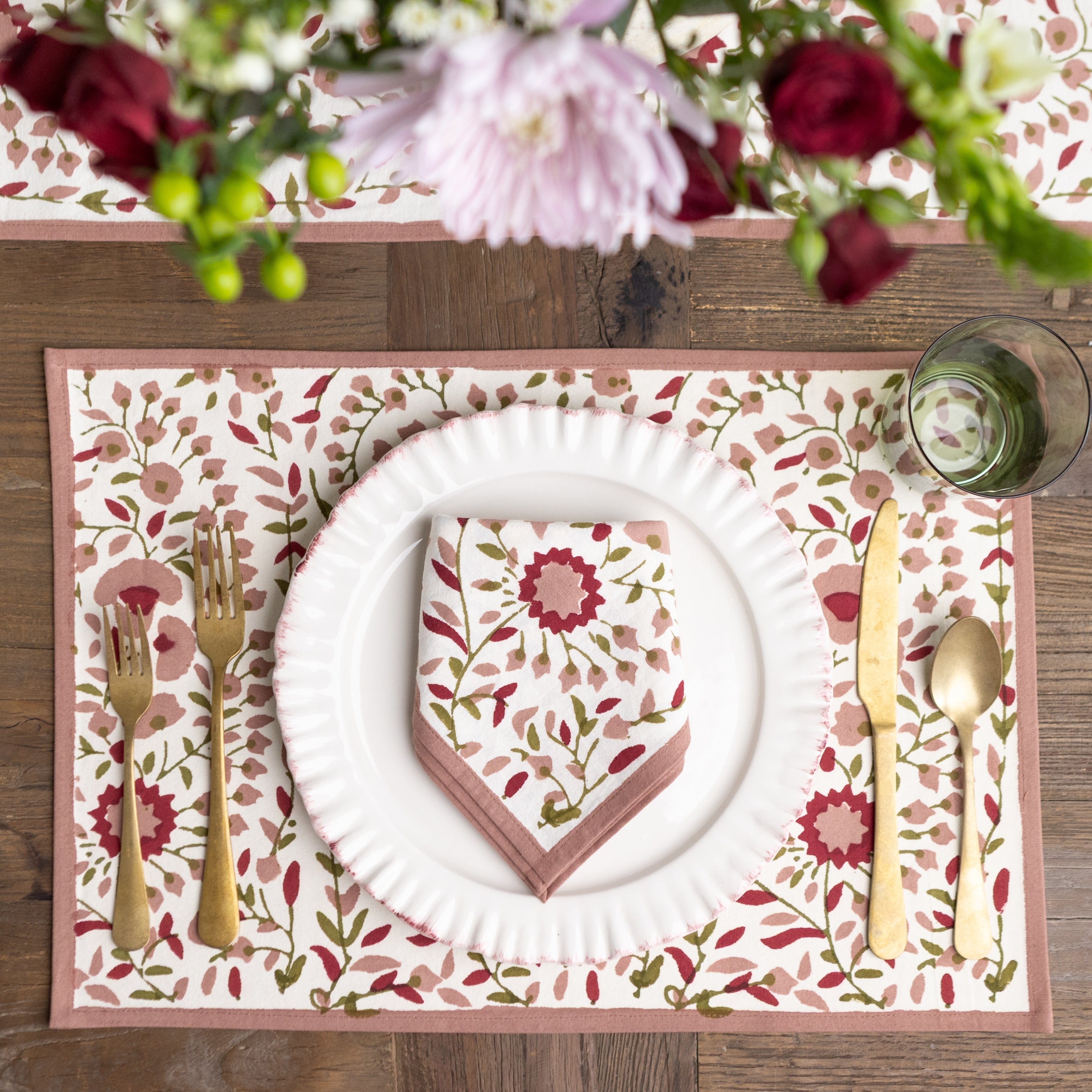 Table setting with floral placemat, white plate, gold cutlery, and pink napkin on a wooden table.