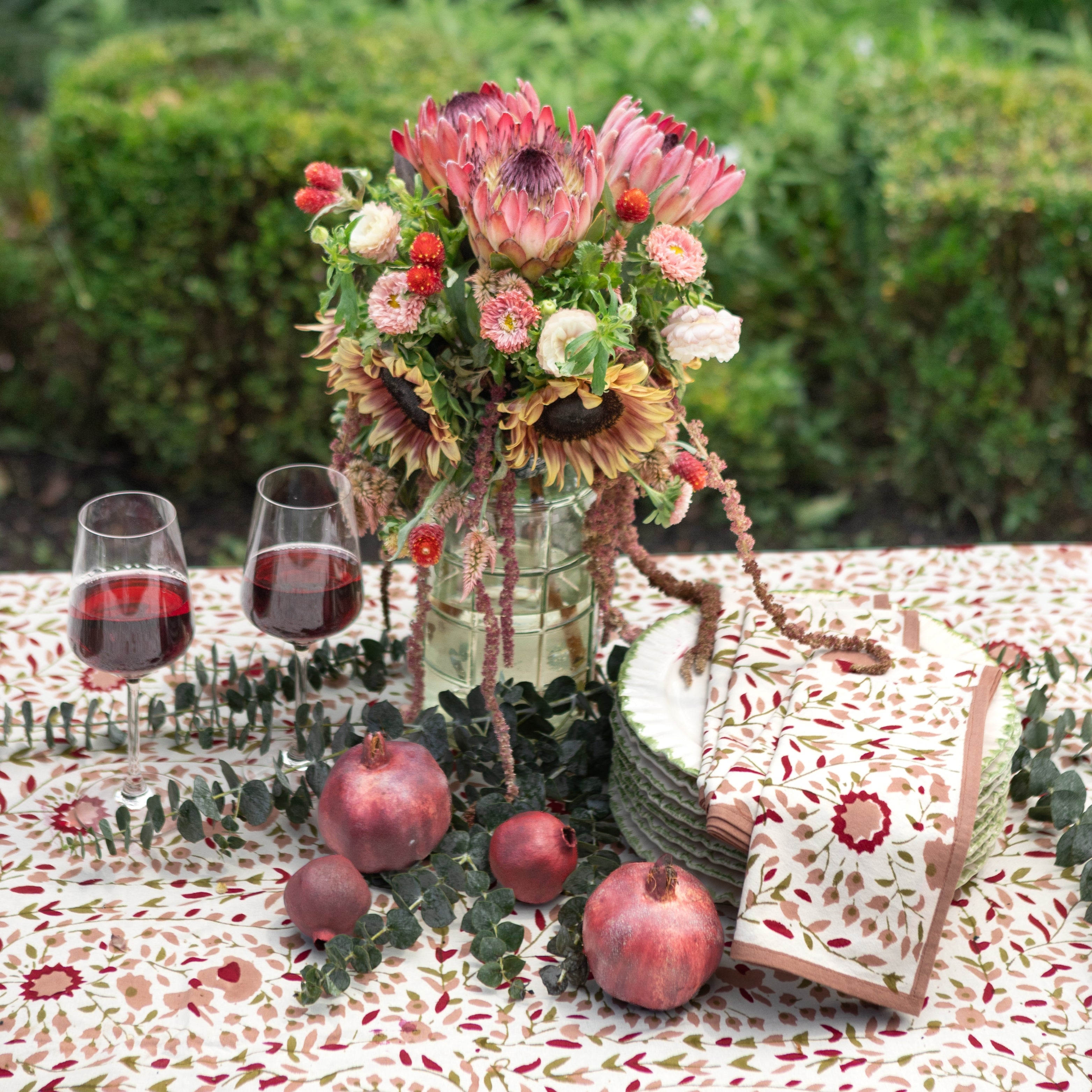 Table setting with flowers, glasses of red wine, pomegranates, and a stack of plates and napkins on a patterned tablecloth.