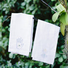 A set of white linen tea towels with blue embroidered designs including a moon and a hand