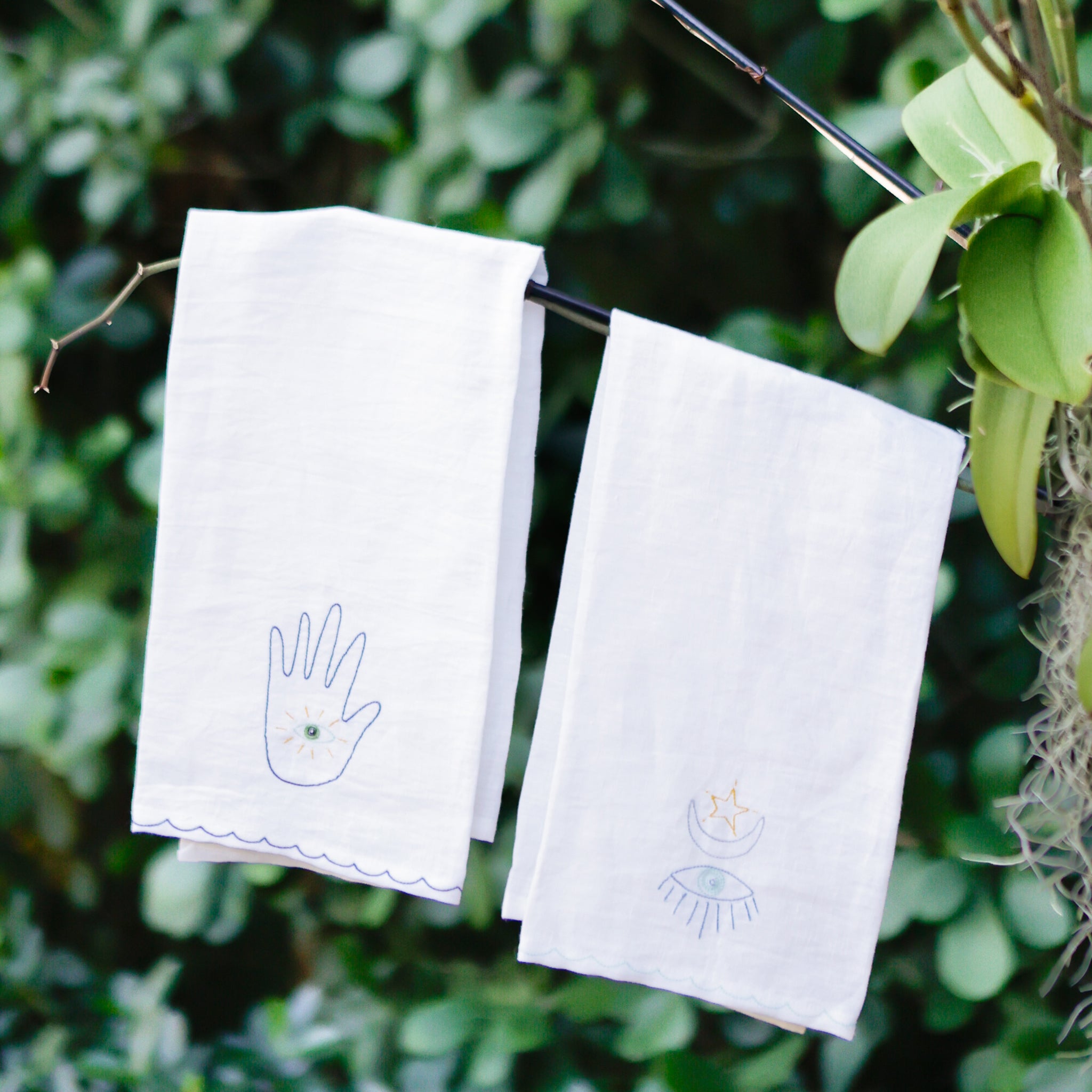 A set of white linen tea towels with blue embroidered designs including a moon and a hand