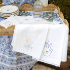 A set of white linen tea towels with blue embroidered designs including a moon and a hand, displayed on a woven tray