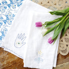 A set of white linen tea towels with blue embroidered designs including a moon and a hand, displayed on a wooden table with tulips