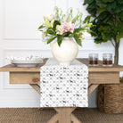 Table setting with a decorative table runner, vase with flowers, and glasses on a wooden table.