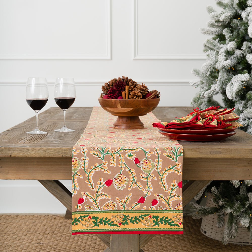 A table runner with a pinecone motif and green and gold border laid out on a table, flanked by two glasses of red wine and a bowl of pinecones, with a Christmas tree in the background.