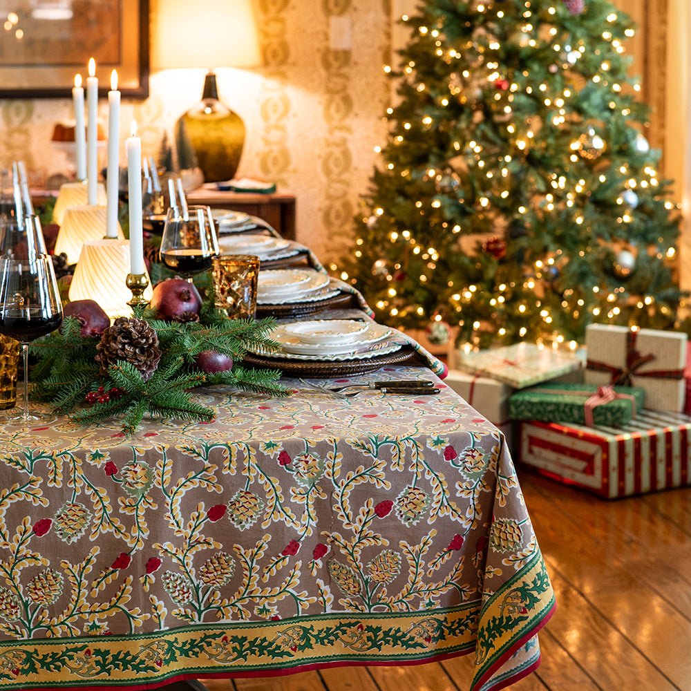 A rectangular tablecloth with a pinecone and foliage pattern, displayed on a table set for dining in doors with a Christmas tree in the background