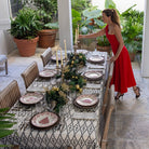 A woman in a red dress is setting a dining table covered with a geometric patterned tablecloth, arranged with plates, cutlery, and candles.