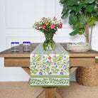 A table runner with a floral pattern displayed on a dining table, flanked by a green vase with flowers and two glasses.