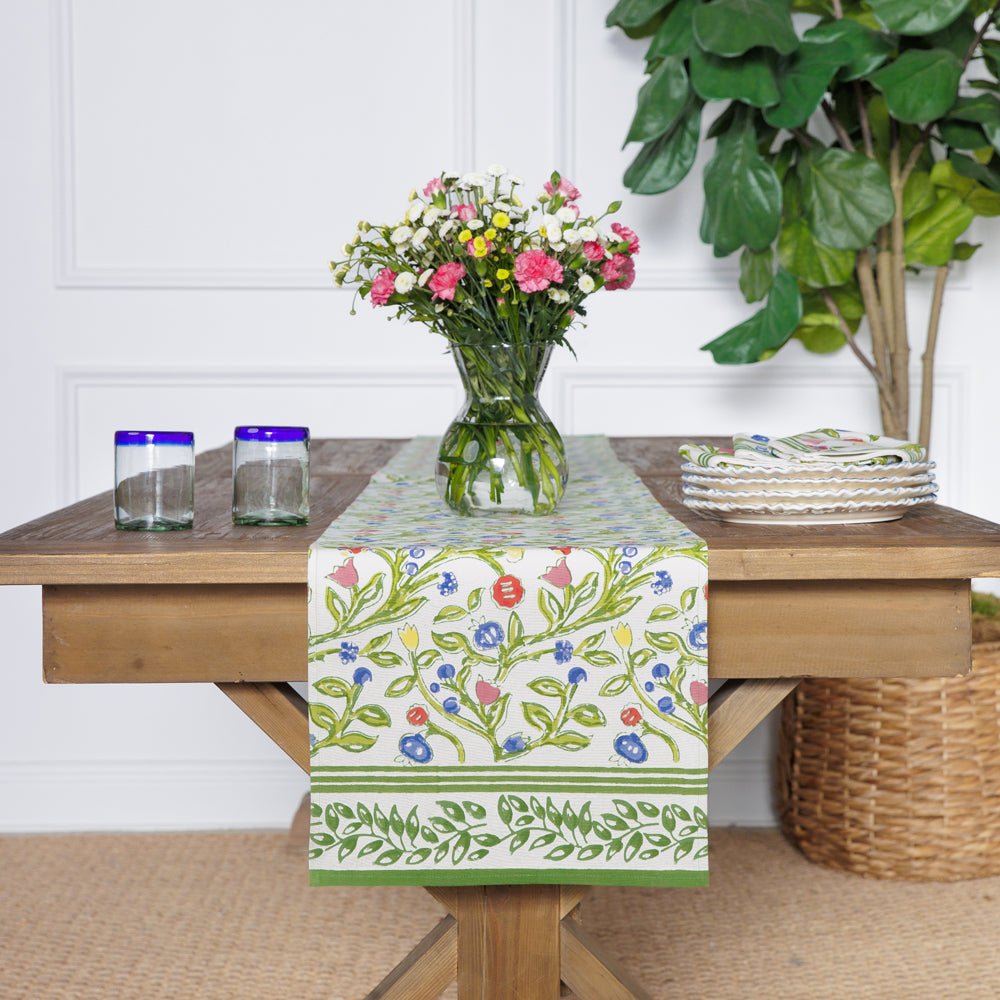 A table runner with a floral pattern displayed on a dining table, flanked by a green vase with flowers and two glasses.
