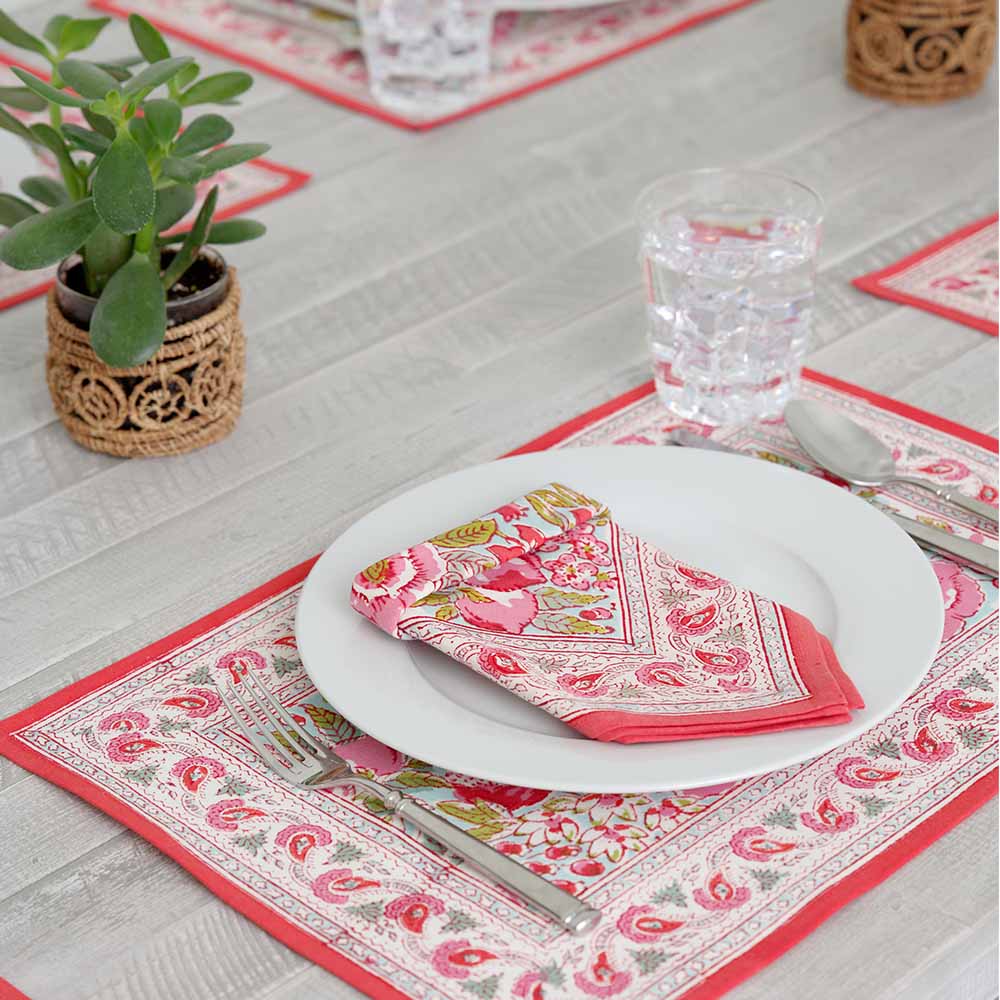 A table setting featuring a rectangular placemat with a floral pattern featuring various shades of pink and green, bordered with a red line.