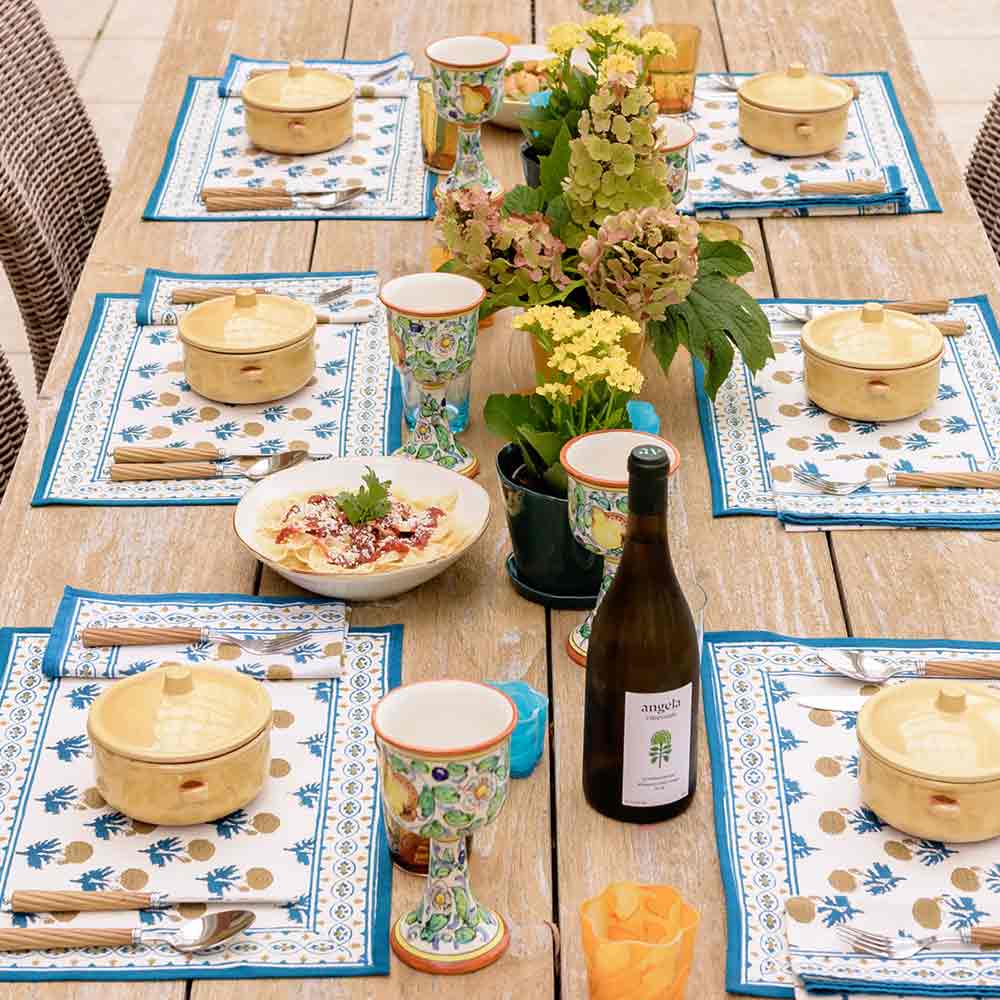Dinner table with blue and yellow floral detailed placemats, flowers, and wine.