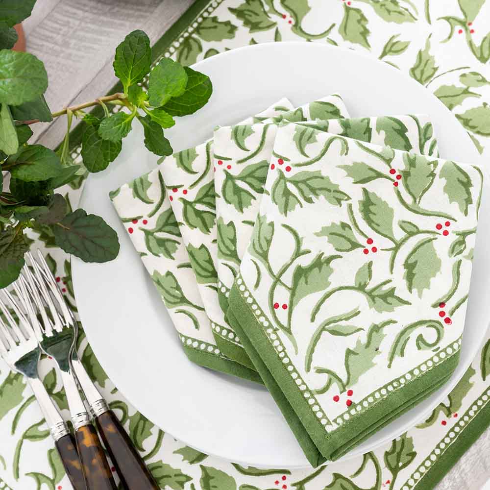 A set of four cotton napkins with a printed green and red holly berry pattern, displayed on a table with cutlery and a plant in the background.