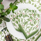 A set of four cotton napkins with a printed green and red holly berry pattern, displayed on a table with cutlery and a plant in the background.