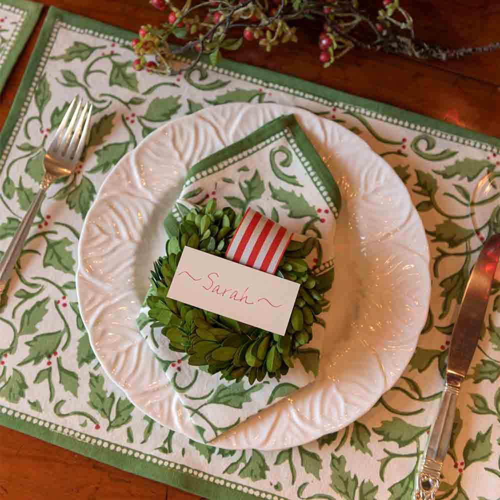 A set of four cotton placemats with a Christmas holly and berries pattern displayed on a table setting alongside a plate and cutlery.