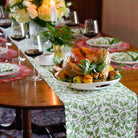 A table runner with a Christmas holly and berries pattern displayed on a table setting with a roasted turkey and holiday decorations.