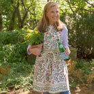 model wearing apron printed with green vines and floral buds in shades of crimson, marigold and deep orange