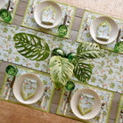 A cotton table runner with a tropical palm and pineapple pattern in light blue and green, displayed on a table with a vase of greenery, plates, and cutlery.